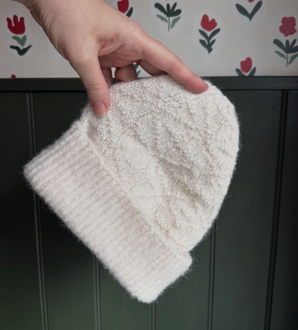 White knitted alpaca wool beanie held by a hand against a floral-patterned wall.