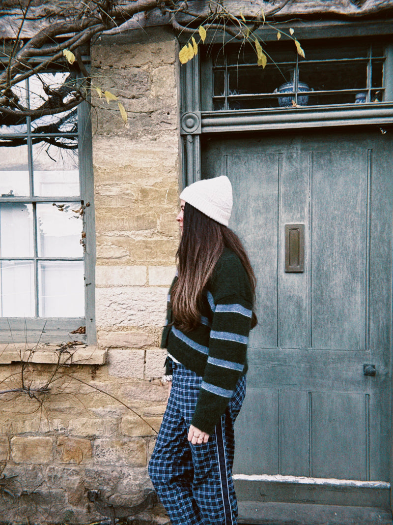 Person wearing a striped sweater and alpaca wool beanie standing in front of a stone building with a wooden door.