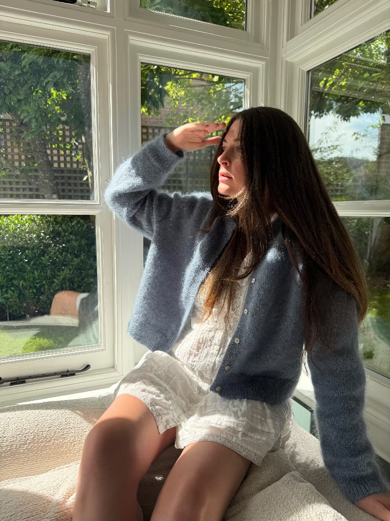 Woman sitting by a window wearing a blue moahir cardigan and white dress, looking out.