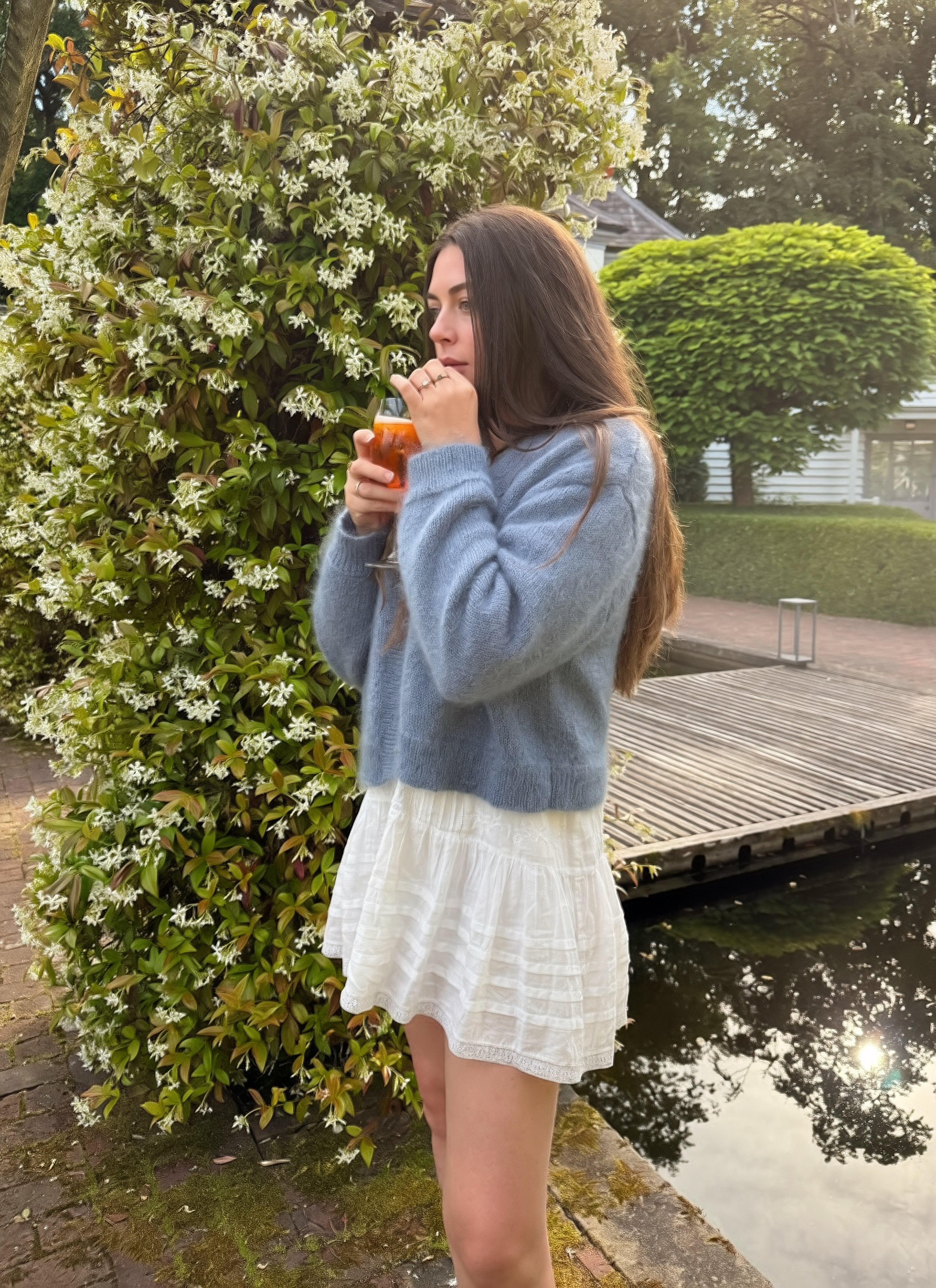Woman in a blue mohair cardigan and white dress standing by a pond with greenery and flowers.