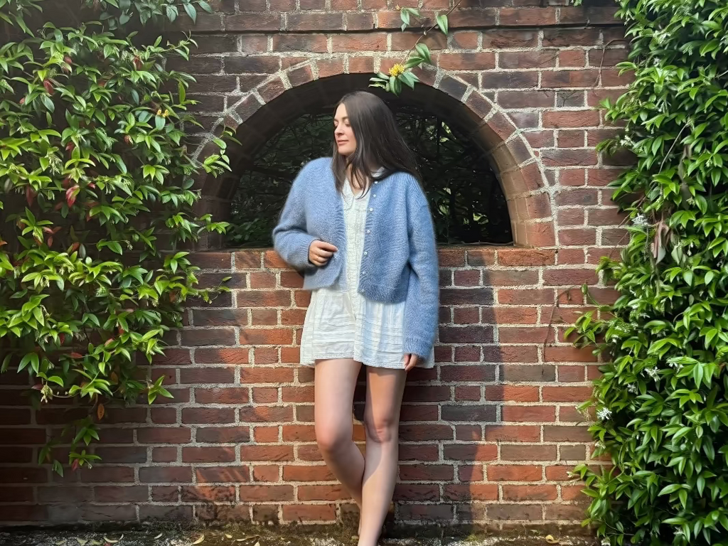 Woman standing in a brick archway in a blue mohair cardigan surrounded by greenery.