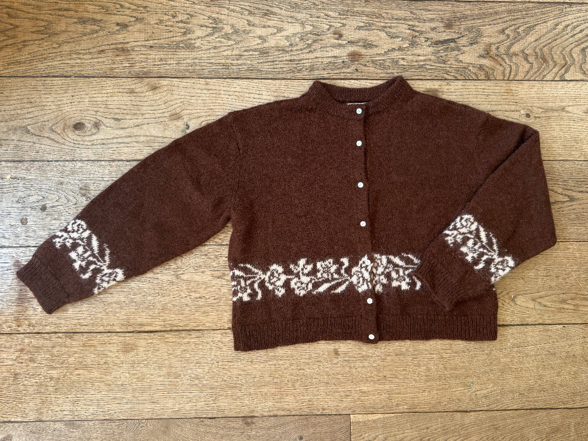 A brown knit cardigan sweater with white floral patterns on the cuffs and hem, laid flat on a light brown wooden plank floor.