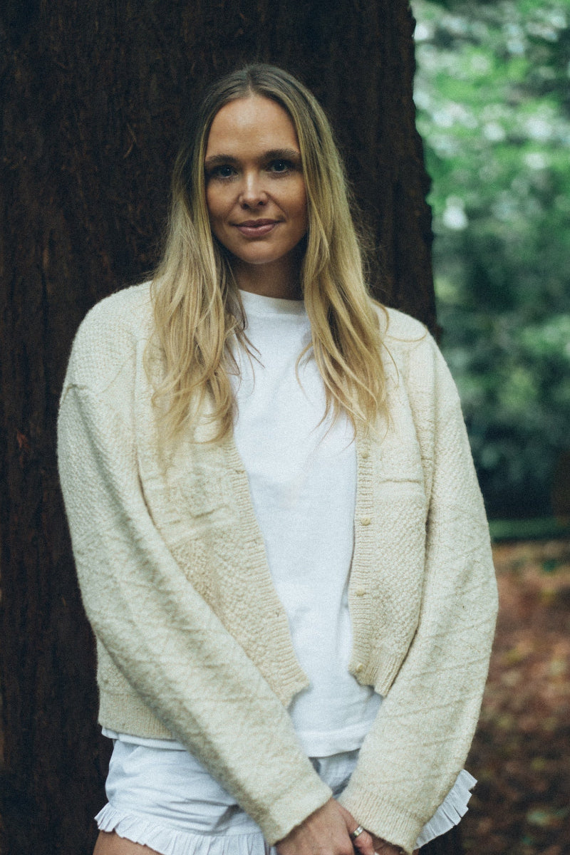 Woman wearing a Cream alpaca wool cardigan over a white shirt and shorts, standing outdoors with trees in the background.