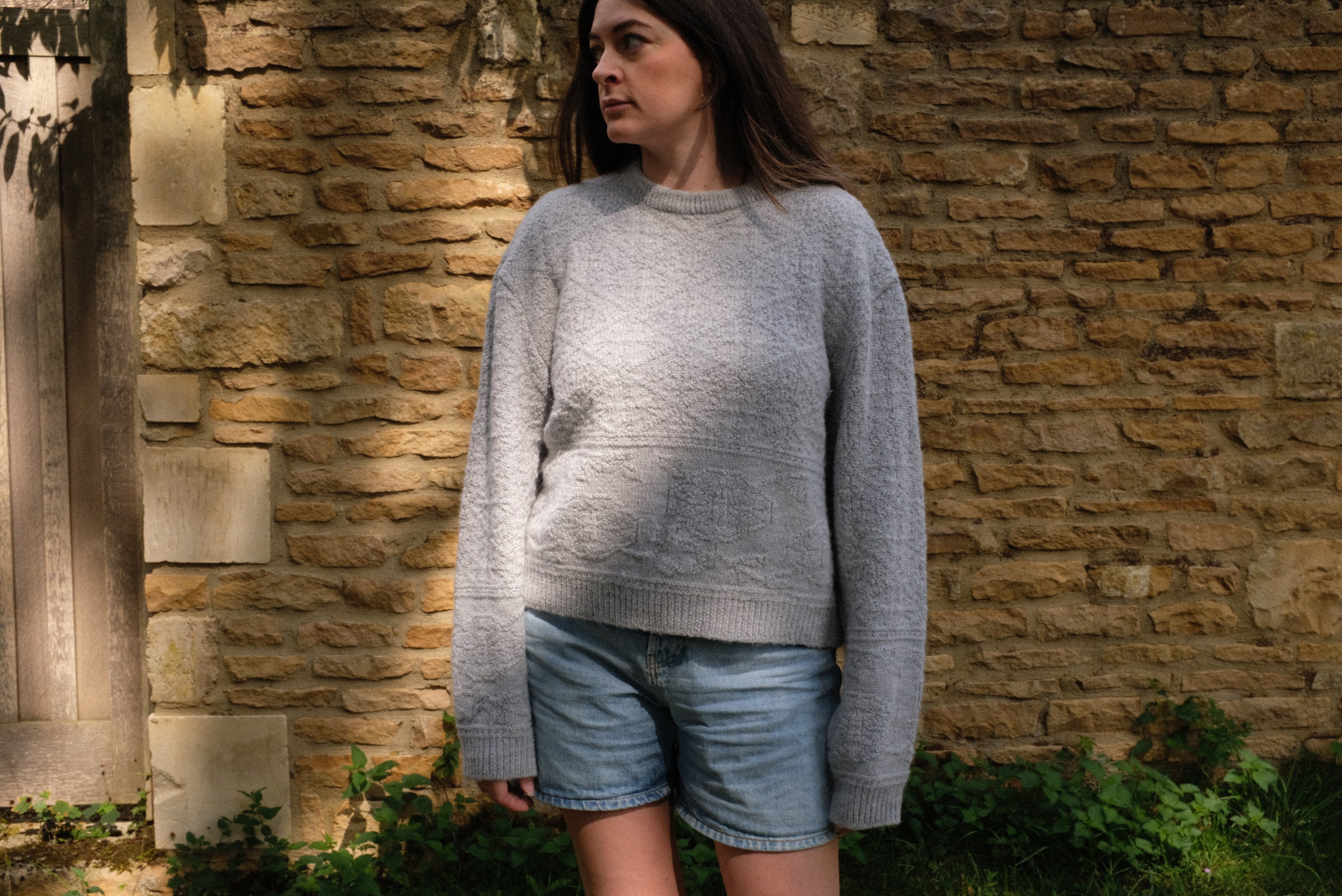 Woman wearing a light grey sustainable knitwear jumper with textured detail, paired with denim shorts.