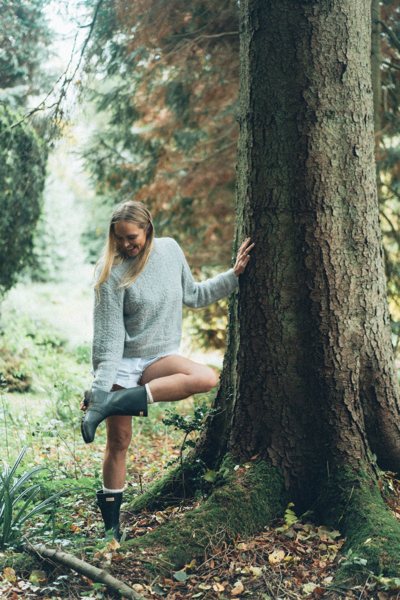 Woman in a forest leaning against a large tree in a Grey textured alpaca wool jumper