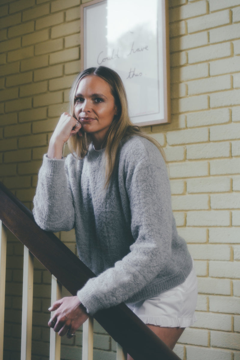Woman in a Grey textured alpaca wool jumper standing against a brick wall.