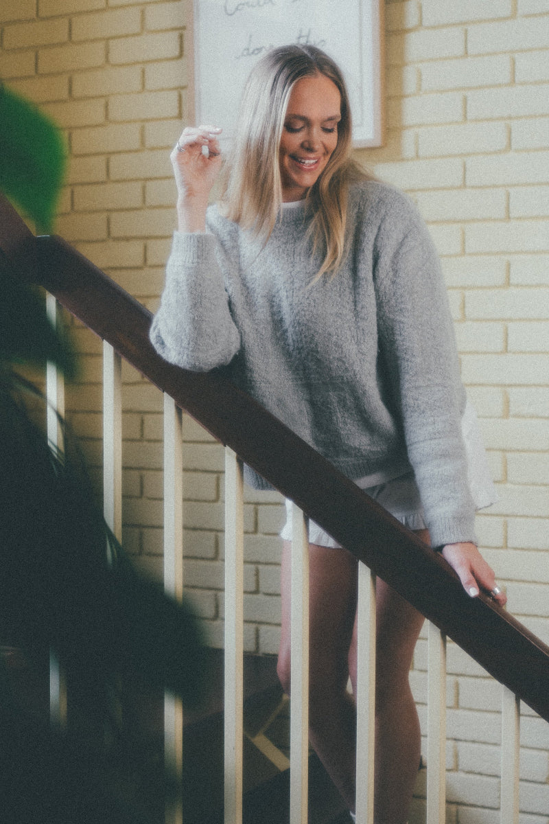 Woman in a Grey textured alpaca wool jumper standing on a staircase with a brick wall background