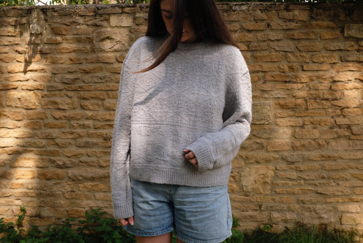 Model wearing a Grey textured alpaca wool jumper in sustainable knitwear paired with denim shorts against a stone wall backdrop.
