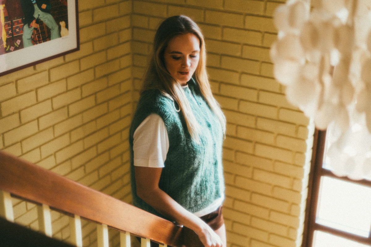 Woman in a green Mohair pointelle knit vest standing in a room with yellow brick walls and a window.
