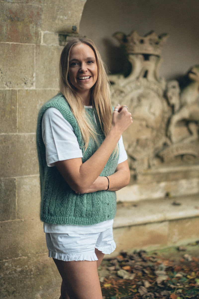 Woman in a green Mohair pointelle knit vest and white shorts standing in front of a stone wall with decorative elements.