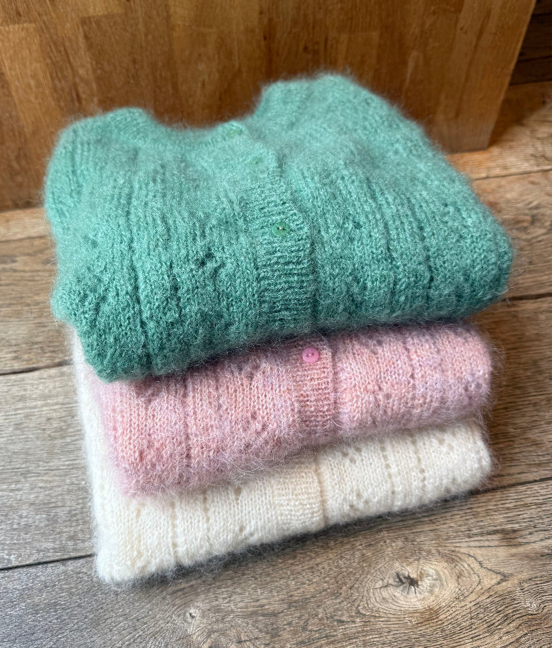 Stack of three folded Mohair pointelle knit vests in green, pink, and white on a wooden surface