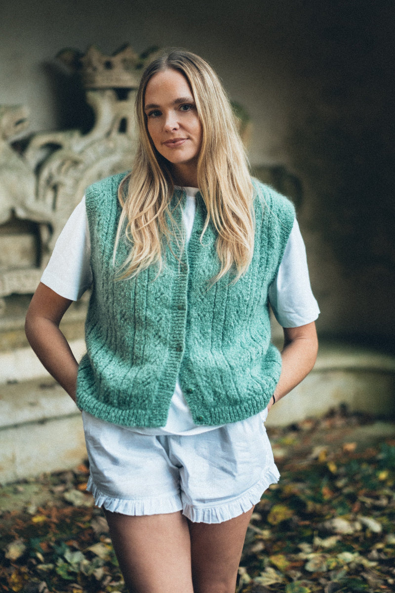 Woman wearing a green Mohair pointelle knit vest over a white shirt and light shorts, standing outdoors.
