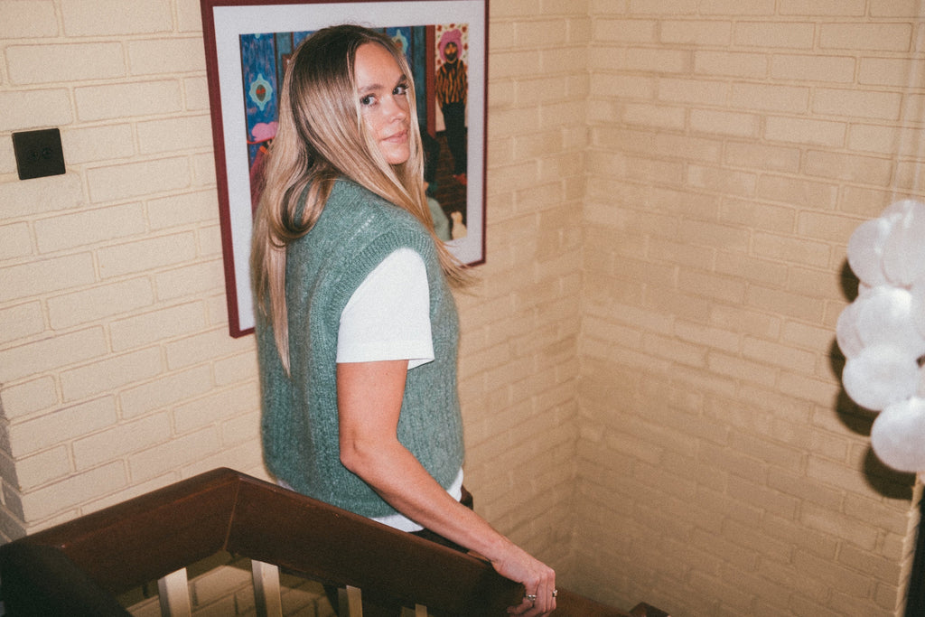 Person wearing a green Mohair pointelle knit vest and white shirt standing in front of a framed picture on a brick wall.