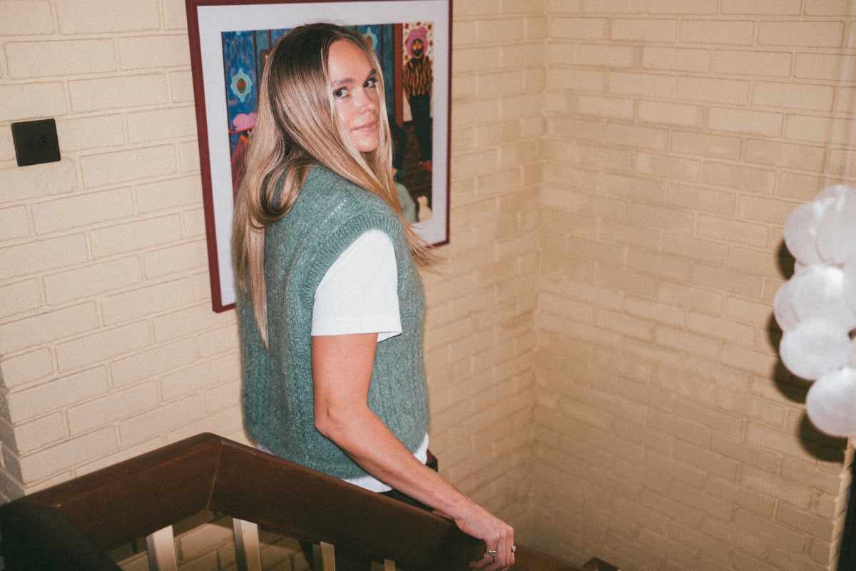 Person wearing a green Mohair pointelle knit vest and white shirt standing in front of a framed picture on a brick wall.